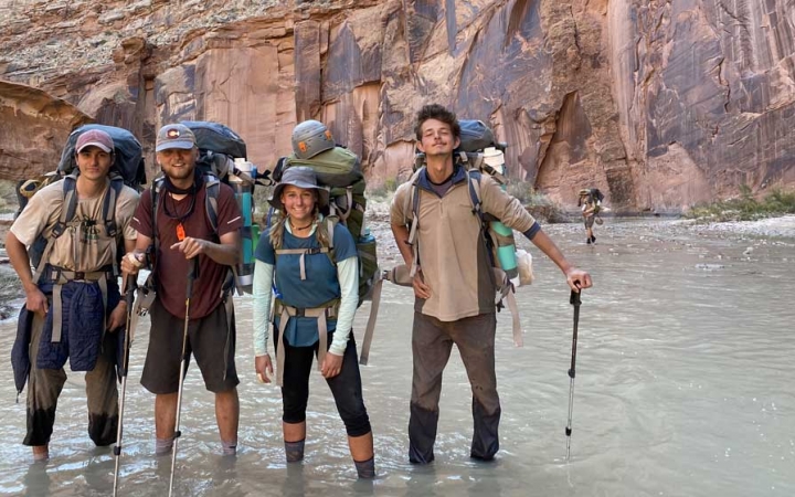 a group of gap year students wearing backpacks stand in a canyon in ankle-deep water